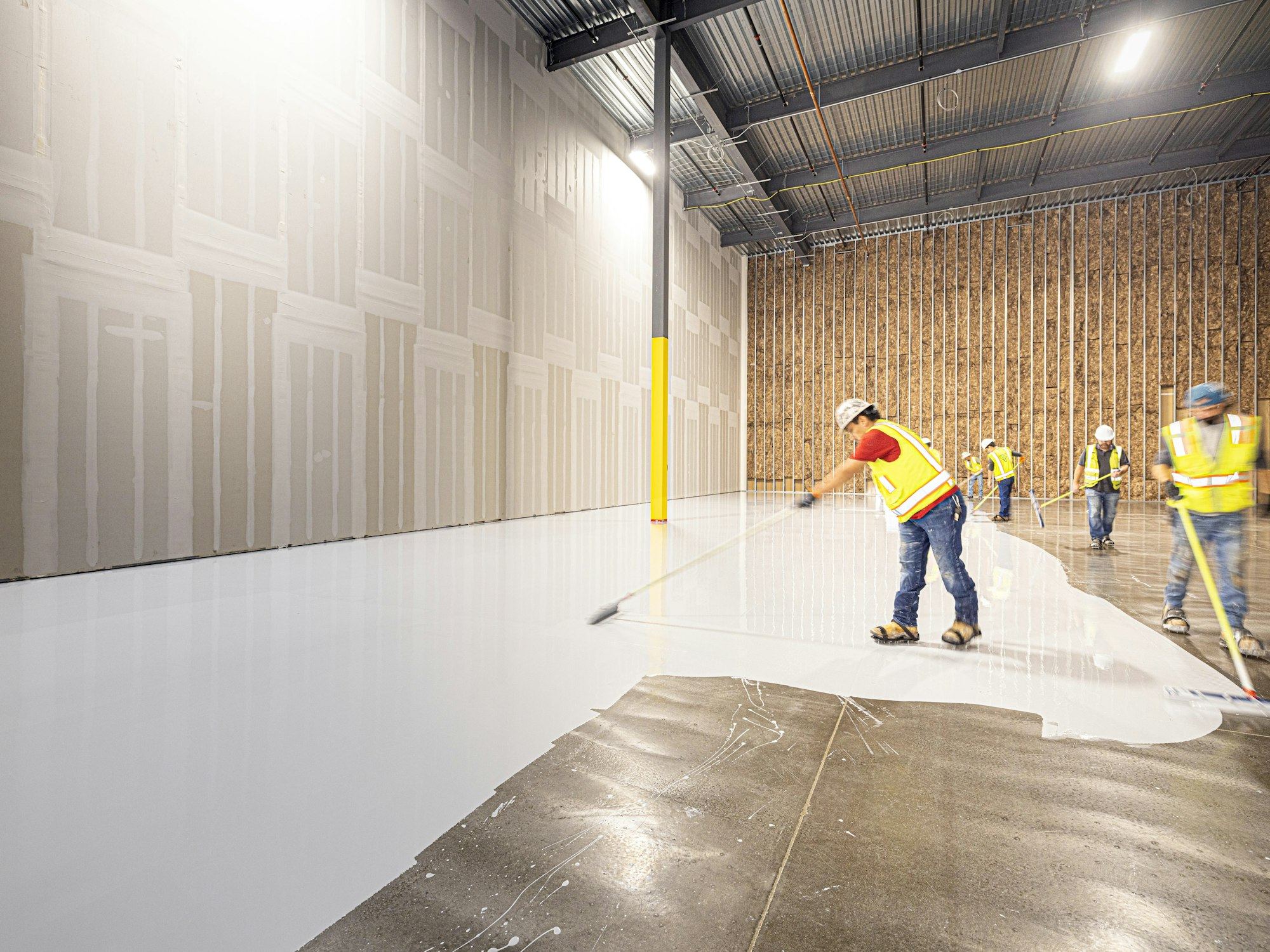 Workers in hi-vis spreading white resin coating across a warehouse floor
